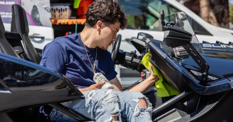A young man in a blue shirt cleans the interior of a black sports car.