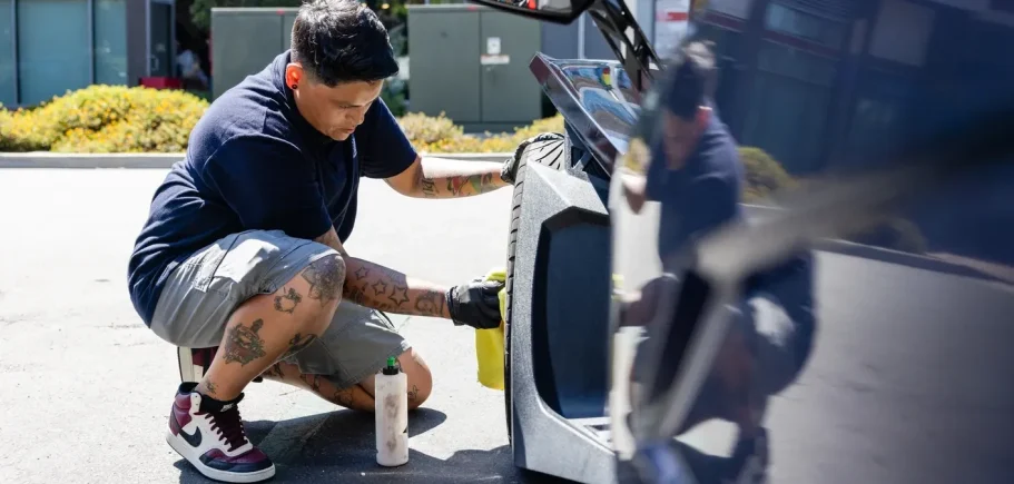 A person wearing a navy shirt and shorts cleans a car's wheel with a spray and cloth.