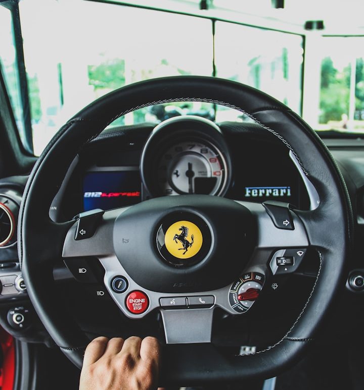 Black steering wheel of a Ferrari with a prominent logo and dashboard instruments visible.